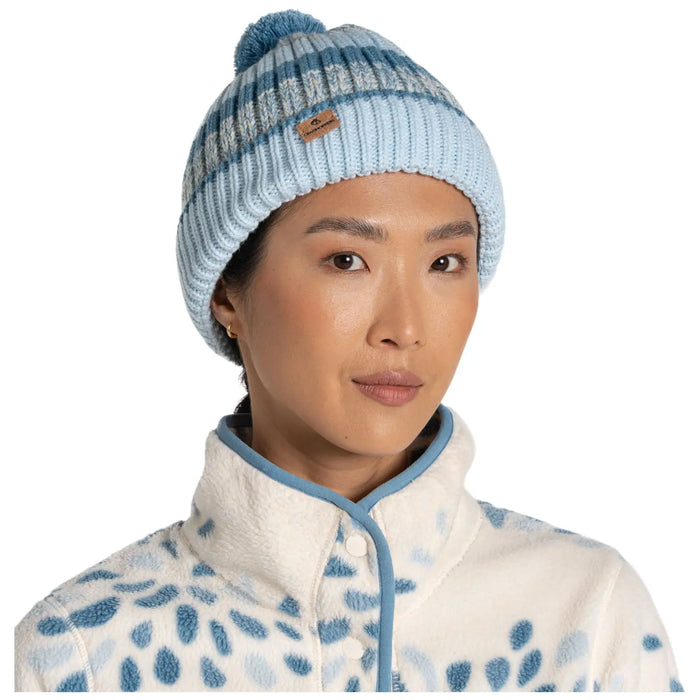 A woman wears a light blue knitted beanie with a pom-pom and stripes while dressed in a cozy white fleece jacket featuring a blue spotted pattern, creating a warm and stylish appearance.