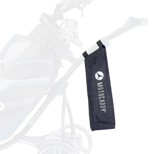 A black towel with the logo "MOTOCADDY" hangs from the handle of a golf trolley placed near a golf bag showcasing a functional accessory for golfers during play.