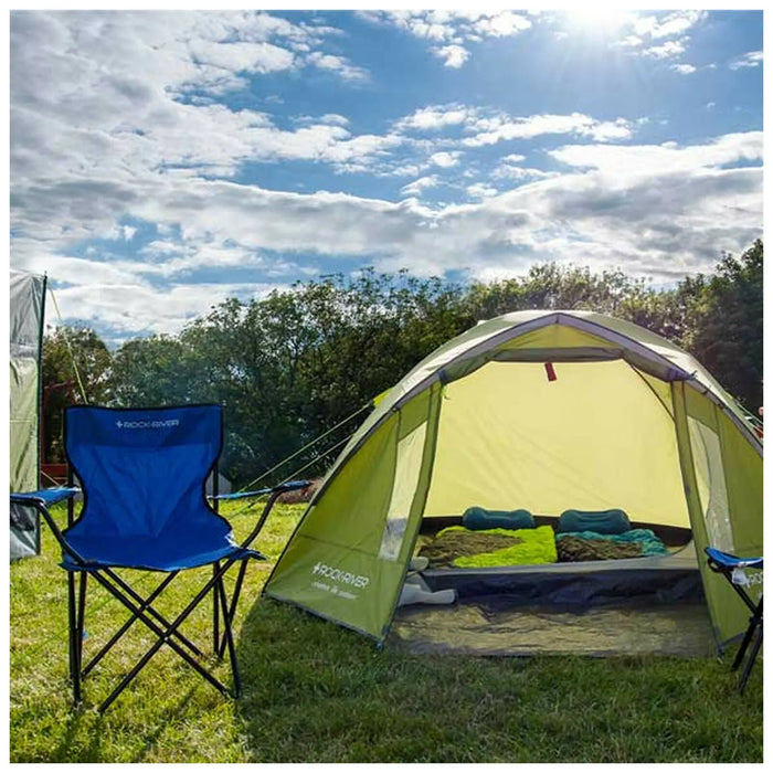 A green tent is set up in a grassy area with a blue camping chair nearby sunlight brightens the scene and clouds float in the sky above surrounding trees create a natural backdrop.