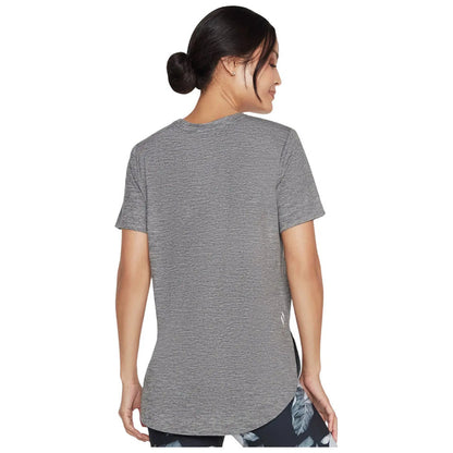 A woman is facing away wearing a grey short-sleeved athletic shirt with a curved hem while in a casual pose against a plain background.