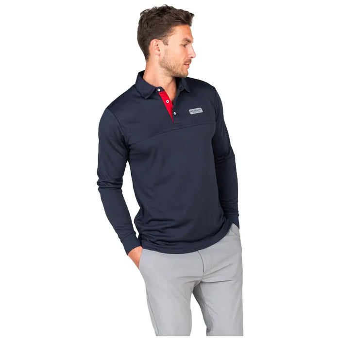 A man wears a dark long-sleeve polo shirt with a red collar and light gray pants standing casually with one hand in his pocket against a plain background.