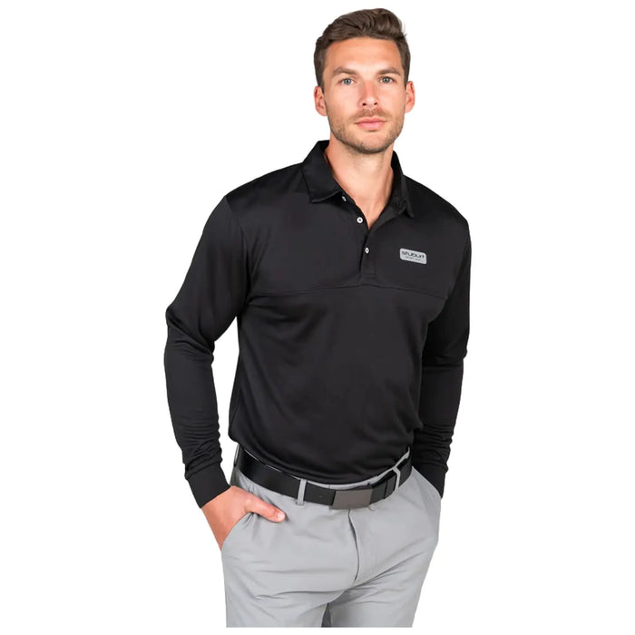A man stands confidently wearing a long-sleeve black polo shirt and gray pants with one hand in his pocket showcasing a casual yet polished look against a plain background.